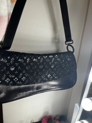 Vintage Svart axelväska från Louis Vuitton - Säljer en svart axelväska från Louis Vuitton med klassiskt monogrammönster i blank finish. Väskan har en avlång, mjukt rundad form och bred axelrem. Materialet är en mix av läder och glansig textil, vilket ger en lyxig känsla och cool look.