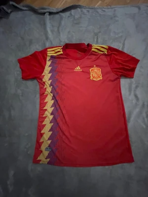 Spaniens matchtröja Adidas röd - Röd matchtröja från Adidas för Spaniens landslag. Tröjan har gula och blå zigzag-detaljer på vänster sida, gula Adidas-ränder på axlarna och landslagets emblem på bröstet. Tillverkad i lätt och ventilerande polyester, perfekt för fotbollsträning eller match.