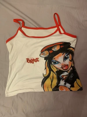 Vit Bratz linnetopp med röd kant - Säljer en vit bratz linne med röd kant. Toppen har tunna axelband och ett stort tryck av en Bratz-karaktär framtill. Perfekt för dig som gillar retro och färgglada motiv. Superskönt material som är stretchigt och enkelt att röra sig i. 