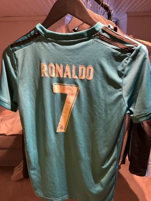 Real Madrid Ronaldo T-shirt - Snygg turkosblå Real Madrid matchtröja från Adidas med Ronaldo och nummer 7 på ryggen. Tröjan har korta ärmar, svarta detaljer på axlarna och grafiskt mönster framtill. Tillverkad i lätt och ventilerande material, perfekt för fotbollsträning.