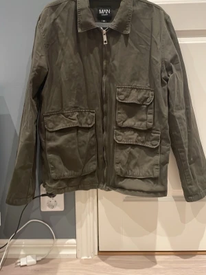 Olivgrön overshirt från MAN - Stilren olivgrön overshirt från MAN med dragkedja framtill och fyra stora fickor med lock. Overshirten har klassisk krage och raka ärmar, perfekt för lager-på-lager. Tillverkad i ett slitstarkt material som ger en schysst look.