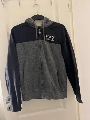 Grå och marinblå hoodie från EA7 Armani - Snygg hoodie från EA7 Emporio Armani i grått och marinblå med dragkedja framtill och justerbar huva med snören. Lite nopprig på insidan annars super bra skick!