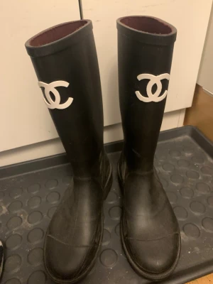 Svarta gummistövlar från Chanel - Säljer ett par svarta höga gummistövlar med  CC loggan framtill.  stövlarna har en stilren, rak form. Perfekta för regniga dagar när du vill hålla stilen. De är endast använda 2-3 gg är som nya.