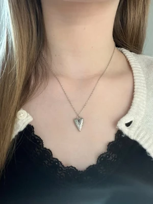 Silverhalsband med hjärthänge - Ett stilrent silverfärgat halsband med ett hjärthänge dekorerat med små pärlor. Kedjan är tunn och elegant, perfekt för att ge en söt touch till din outfit. Levereras i en fin ask. Rostfritt stål och handgjort 