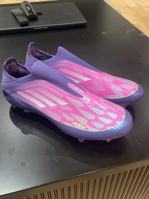 Adidas X Speedflow+ lila/rosa fotbollsskor - Säljer ett par Adidas X Speedflow+ fotbollsskor i lila och rosa med silverdetaljer. Skorna har en strömlinjeformad design utan snörning, tillverkade i syntetmaterial för lätt vikt och bra grepp på planen. Perfekta för dig som vill sticka ut på fotbollsplanen.