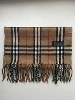 Burberrys beige rutig halsduk ull - Klassisk beige halsduk från Burberrys i 100% lammull med ikoniskt rutmönster i svart, vitt och rött. Halsduken har fransar i ändarna och är tillverkad i England. Perfekt accessoar för att lyfta din outfit.