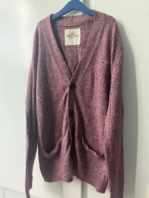 Vinröd kofta från Hollister - Mysig vinröd kofta från Hollister med två knappar framtill och två fickor. Koftan har en avslappnad passform, V-ringning och diskret logga broderad på bröstet. Perfekt att slänga på sig över en t-shirt för en chill look.