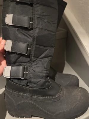 Ridvinterstövlar från Cavaliero stl 36 - Säljer ett par svarta vinterstövlar från Covaligo. De har högt skaft i vadderat tyg, tre breda kardborreband och slitstark gummisula med grovt mönster. Perfekta för kalla dagar och snö, med vattentålig nederdel och robust design.