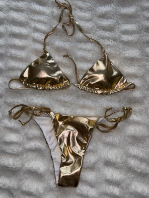 Guldig bikini med knytband - Snygg guld bikini med trekantsöverdel och knyt i både sidor och nacke. Glansigt material som verkligen sticker ut på stranden. Underdelen har knyt i sidorna för justerbar passform. Perfekt för sommarens alla bad. Köpt på amazon och aldrig använd. Storlek S