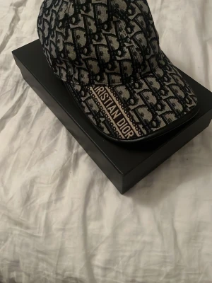 Monogram keps från Christian Dior - Säljer en snygg keps från Christian Dior med ikoniskt monogrammönster i svart och grått. Kepsen har böjd skärm och broderad logga på framsidan. Tillverkad i textilmaterial som känns lyxigt och trendigt. Perfekt accessoar för dig som gillar designerplagg.