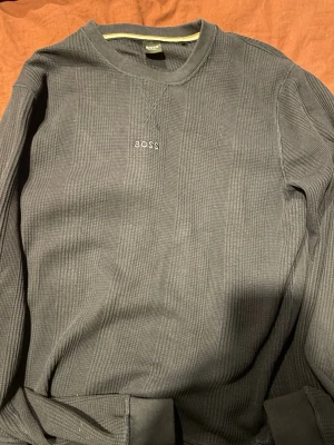 Mörkblå loungewear-tröja från BOSS - Mysig mörkgrå loungewear-tröja från BOSS Homewear med diskret ribbad struktur och broderad logga på bröstet. Tröjan har rund halsringning och en gul/vit rand på insidan av kragen. Perfekt för chill dagar hemma.