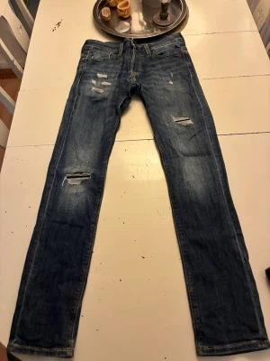 Blå slitna jeans med raka ben - Säljer ett par blå jeans med slitningar och hål på låren och knäna. Jeansen har klassisk femficksmodell, normal midja och raka ben. Materialet är denim i bomull och de har en cool, avslappnad look. Behöver säga det att de är lite sönder i skrevet men det är en relativ enkel fix men så ni vet.