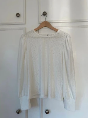 Vit broderad blus med puffärm - Söt vit blus med broderade detaljer och rund halsringning. Blusen har puffiga långärmar med breda manschetter och ett romantiskt hålmönster i tyget. Perfekt för dig som gillar en feminin och stilren look.
