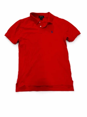 Röd pikétröja från Polo Ralph Lauren - Klassisk röd pikétröja från Polo Ralph Lauren med blå logga broderad på bröstet. Tröjan har krage, knappar framtill och korta ärmar. Perfekt för en stilren och sportig look.