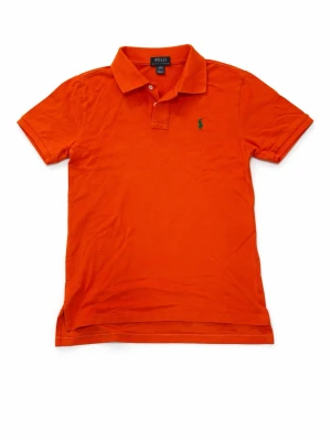 Orange pikétröja från Polo Ralph Lauren - Klassisk orange pikétröja från Polo Ralph Lauren med krage, korta ärmar och liten grön broderad logga på bröstet. Perfekt för en avslappnad och stilren look.