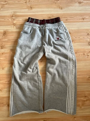 Grå mjukisbyxor Alfric Eden - Grå mjukisbyxor med breda ben, vita ränder längs sidorna och elastisk midja i röd- och svartmönstrat tyg. Broderad röd hjärtlogga och text på vänster sida. Mjukt material och fickor framtill.