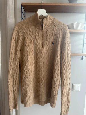 Polo Ralph Lauren, storlek M - Säljer en beige kabelstickad tröja från Polo Ralph Lauren med halv dragkedja och hög krage. Tröjan har det klassiska broderade logotypen på bröstet och är gjord i mjukt material. 90% ull, 10% Kashmir. Självklart äkta, med QR kod. Superfin, inget att anmärka på. 