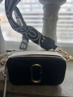 Marc Jacobs Snapshot svart/vit väska - Säljer en Marc Jacobs Snapshot handväska i svart och vitt med guldfärgade detaljer och bred axelrem med logotyp. Väskan har två dragkedjor upptill och är tillverkad i läder med canvasrem. Perfekt för dig som vill ha en trendig och praktisk accessoar.  PRIS KAN DISKUTERAS 