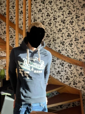 Mörkblå Superdry hoodie med tryck - Snygg mörkblå hoodie från Superdry med vitt och mintgrönt tryck framtill. Hoodien har en stor huva med vita dragsnören och en klassisk magficka. Perfekt för en avslappnad streetwear-look.
