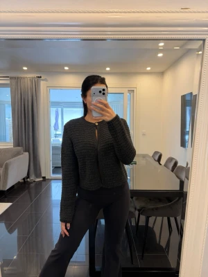 Svart stickad kofta från Zara - Mörkgrå stickad kofta från Zara med rund halsringning och en dekorativ guldig knapp upptill. Koftan har ett grovt stickat mönster, raka ärmar och en något boxig passform. Perfekt att slänga över en topp för en snygg lager-på-lager-look.