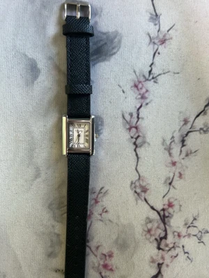 Svart rektangulär klocka med läderband - Stilren klocka med svart läderband och rektangulärt silverfärgat ur. Urtavlan är vit med romerska siffror och svarta visare. Perfekt accessoar för en klassisk look. Batteri behövs bytas men det är enkelt