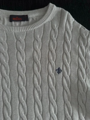 Vit kabelstickad tröja Morris - Vit kabelstickad tröja från Morris med rund halsringning och blå broderad logga på bröstet. Tröjan har långa ärmar och ribbade muddar vid ärmslut och nederkant. Perfekt för en klassisk och stilren look⭐️