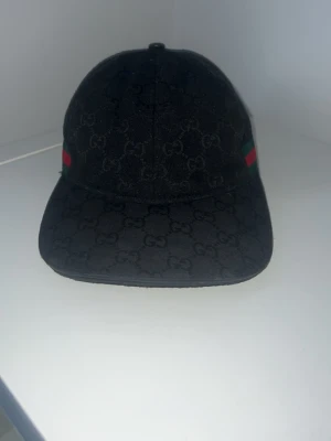Svart Gucci keps med GG-mönster - Snygg svart keps från Gucci med klassiskt GG-monogram över hela kepsen och detaljer i rött och grönt på sidorna. Justerbar rem baktill och tillverkad i ett slitstarkt tygmaterial. Perfekt för dig som vill ha en exklusiv och trendig accessoar.