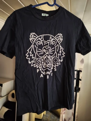 Svart K t-shirt med tigertryck - Snygg svart t-shirt från Kmed ikoniskt vitt tigertryck och texten 'K Paris' på bröstet. Klassisk rund hals och korta ärmar. Tillverkad i mjuk bomull som känns skön mot huden. Perfekt för dig som vill ha en statement-tee i garderoben.