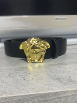 Svart skärp med guldspänne från Versace - Stilrent svart skärp från Versace med ikoniskt Medusa-spänne i guld. Skärpet är gjort i äkta skinn och har en klassisk, tidlös design som lyfter vilken outfit som helst.