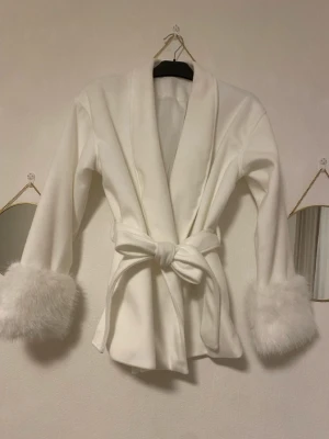 Vit pälsväst med knytskärp - Stilren vit väst med fluffiga fuskpälsdetaljer vid ärmsluten och knytskärp i midjan. Västen har en öppen design med bred krage och ger en lyxig känsla. Perfekt för att lyfta din outfit med en trendig touch.