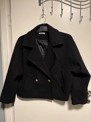 Svart skepparkavaj med guldknappar - Stilren svart skepparkavaj med bred krage och dubbla guldknappar framtill. Jackan har två fickor och är fodrad på insidan. Perfekt för dig som vill ha en klassisk look med modern touch. 