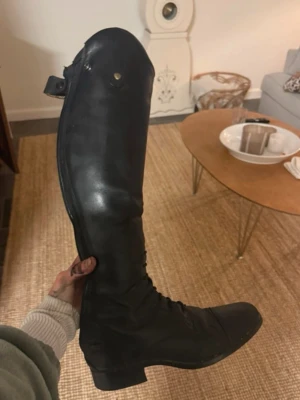 Svarta höga ridstövlar i skinn - Stilrena svarta ridstövlar i skinn med snörning framtill och hög skaft. Perfekta för dig som gillar klassisk ridstil. Snygg och tidlös design som passar till många outfits.storlek 37,5
