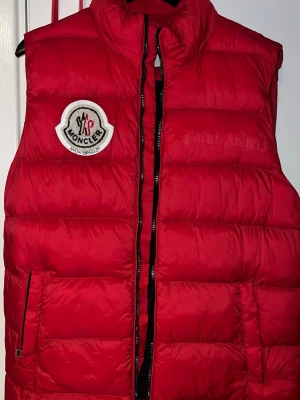 Röd dunväst från Moncler - Riktigt snygg röd dunväst från Moncler med klassisk logga på bröstet och svarta dragkedjor. Västen har en quiltad design och två fickor framtill. Insidan har ett färgglatt Moncler-tryck. Perfekt för lager-på-lager-stil.