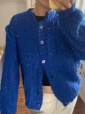 Kornblå stickad kofta med knappar - Säljer en kornblå stickad kofta med stora lila knappar framtill. Koftan har en lös passform, ribbstickade muddar och ett luftigt, hålmönstrat material. Perfekt för lager-på-lager och ger en cool, avslappnad vibe.