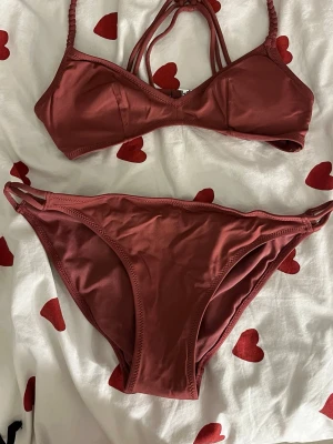 Mörkröd bikini med flätade band - Snygg mörkröd bikini med flätade axelband och flera smala band i ryggen. Överdelen har en enkel, stilren design och underdelen har dubbla sidoband för extra detalj. Perfekt för stranden eller poolen.
