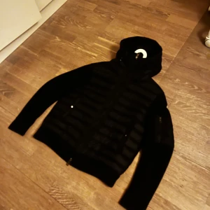 Svart dunväst med huva från Moncler - Snygg svart dunväst från Moncler med huva och quiltad front. Västen har dragkedja, två fickor med dragkedja och Moncler-logga på bröstet. Ärmarna är i mjukt material som kontrasterar mot den glansiga dunfronten. Perfekt för lager på lager och streetwear.