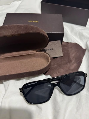 Tom Ford glasögon - Snygga solglasögon från Tom Ford med tillhörande brun sammetsask, putsduk och originalförpackning. Lyxig känsla med gulddetaljer på asken och klassisk design. Perfekt accessoar för att lyfta din stil.