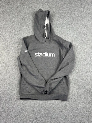 Grå hoodie från Stadium med tryck - Mörkgrå hoodie från Stadium med vitt tryck på bröstet och logga på ärmen. Klassisk känguruficka framtill, ribbade muddar och justerbar huva med snören. Perfekt för en avslappnad och sportig stil.