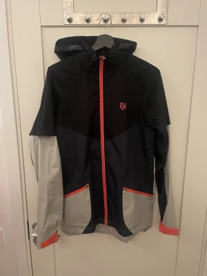 Footkorner vindjacka - Snygg vindjacka från footkorner köpt i paris i svart och grått med röda detaljer. Jackan har huva, hel dragkedja framtill och två fickor med röda dragkedjor. Materialet är tunt och vindtätt, perfekt för blåsiga dagar. Footkorner-logga på bröstet och ryggen.