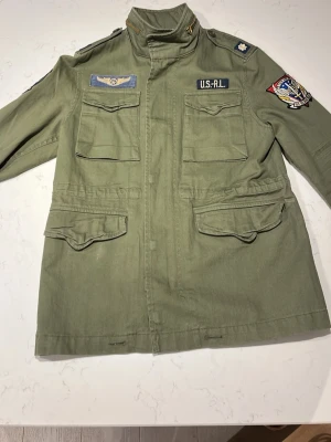 Ralph Lauren Field Jacket - Militärgrön Field Jacket från Ralph Lauren med flera coola patches och bröstfickor. Jackan har hög krage, fyra fickor framtill och detaljerade broderade märken på ärmen och bröstet. Perfekt för dig som gillar utility-stil.