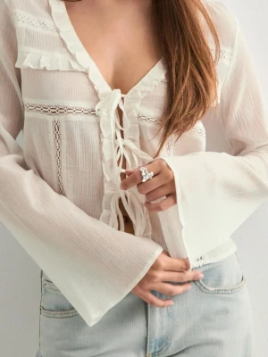 Vit blus med volanger och knytning - Romantisk vit blus med långa ärmar, volangdetaljer och knytning framtill. Blusen har spetsinslag och lätt transparent tyg som ger en luftig känsla. Perfekt att styla med jeans eller kjol för en trendig look.