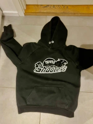 Svart Trapstar Shooters hoodie - Snygg svart hoodie från Trapstar med stort broderat Shooters-motiv på bröstet i vitt och svart. Klassisk känguruficka framtill och huva med dragsko. Perfekt för dig som gillar streetwear och vill sticka ut med en unik design.
