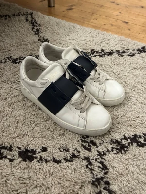 Valentino Garavani Open sneakers - Säljer ett par vita Valentino Garavani Open sneakers med bred marinblå läderdetalj över snörningen. Skorna har rund tå, platt sula och klassiska snören. Tillverkade i skinn och har diskret Valentino-logga på plösen. Perfekta för dig som vill ha en clean men lyxig look.