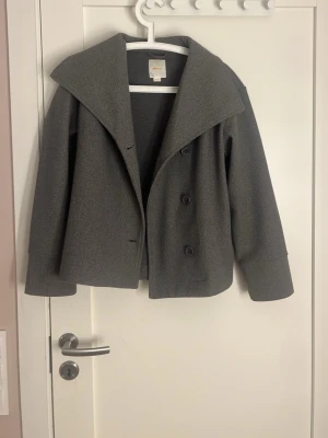 Grå trenchcoat  - Stilren grå kort rock från Gina Tricot med bred krage och dubbelknäppning framtill. Tillverkad i ullblandning och har en boxig, modern passform. Perfekt för dig som gillar minimalistisk och trendig stil.