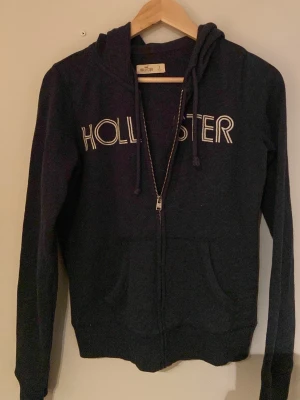 Mörkblå zip-hoodie från Hollister - Mörkblå hoodie med dragkedja från Hollister. Stor logga i vitt framtill, två fickor och justerbar huva med snören. Mjuk bomullsblandning och ribbade muddar vid ärmslut och nederkant. Perfekt för en avslappnad och stilren look.