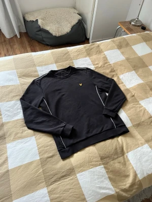Svart sweatshirt från Lyle & Scott - Svart sweatshirt från Lyle & Scott med klassisk rund hals och gul logga på bröstet. Tröjan har vita detaljer längs sidorna och är långärmad med ribbade muddar. Perfekt för en avslappnad och stilren look.