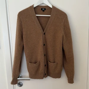 Beige Cardigan från Uniqlo  - Snygg beige cardigan från Uniqlo i mjuk ull. Modellen har v-ringning, svarta knappar framtill och två praktiska fickor. Perfekt för lager-på-lager och passar dig som gillar enkel och stilren design. Den är använd men har mycket liv kvar.