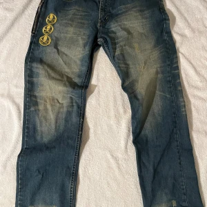 Vintage Levi's jeans med patchar och slitningar - Blå Levi's jeans med coola slitningar och ljusa partier, samt tre gula dödskallepatchar på vänster lår. Klassiska bakfickor med kontrastsömmar och röd Levi's-tagg. Jeansen har rak-baggy passform och är tillverkade i robust denim.