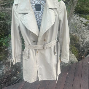 Beige trenchcoat från Max Mara Weekend - Stilren beige trenchcoat från Max Mara Weekend med klassisk dubbelknäppning, bälte i midjan och snygga detaljer vid ärmslut. Jackan har ett blommigt foder och stora slag. Perfekt för dig som vill ha en tidlös och trendig look.