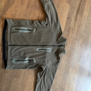 Grå / beige fleecejacka från Arc'teryx - Säljer en grå fleecejacka från Arc'teryx med hög krage och hel dragkedja. Jackan har flera praktiska fickor med dragkedja och diskret logga på bröstet. Materialet är mjukt och strukturerat, perfekt för lager-på-lager under kyliga dagar. This is a real vintage, produced before qr codes. Been in nice adventures but still in very good shape